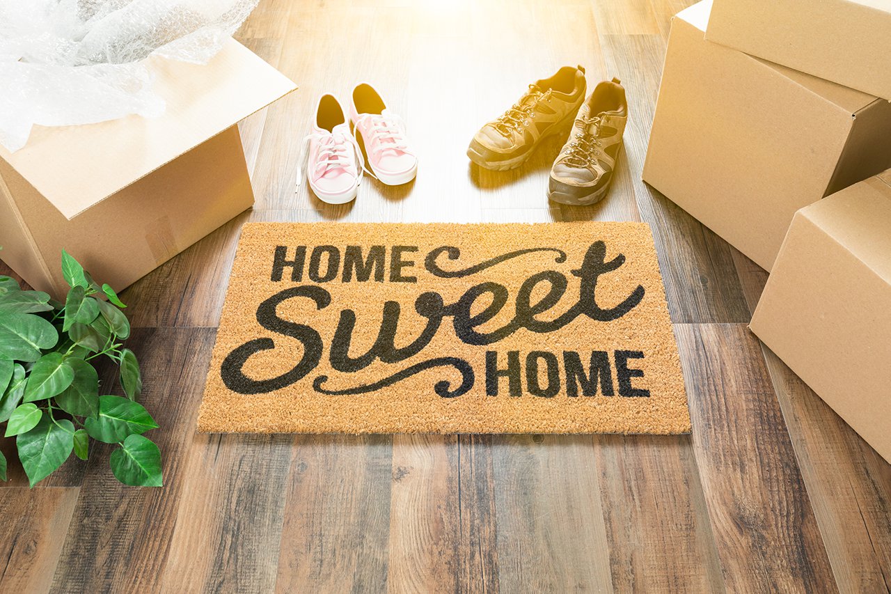Home-Sweet-Home-Welcome-Mat,-Moving-Boxes,-Women-and-Male-Shoes-and-Plant-on-Hard-Wood-Floors